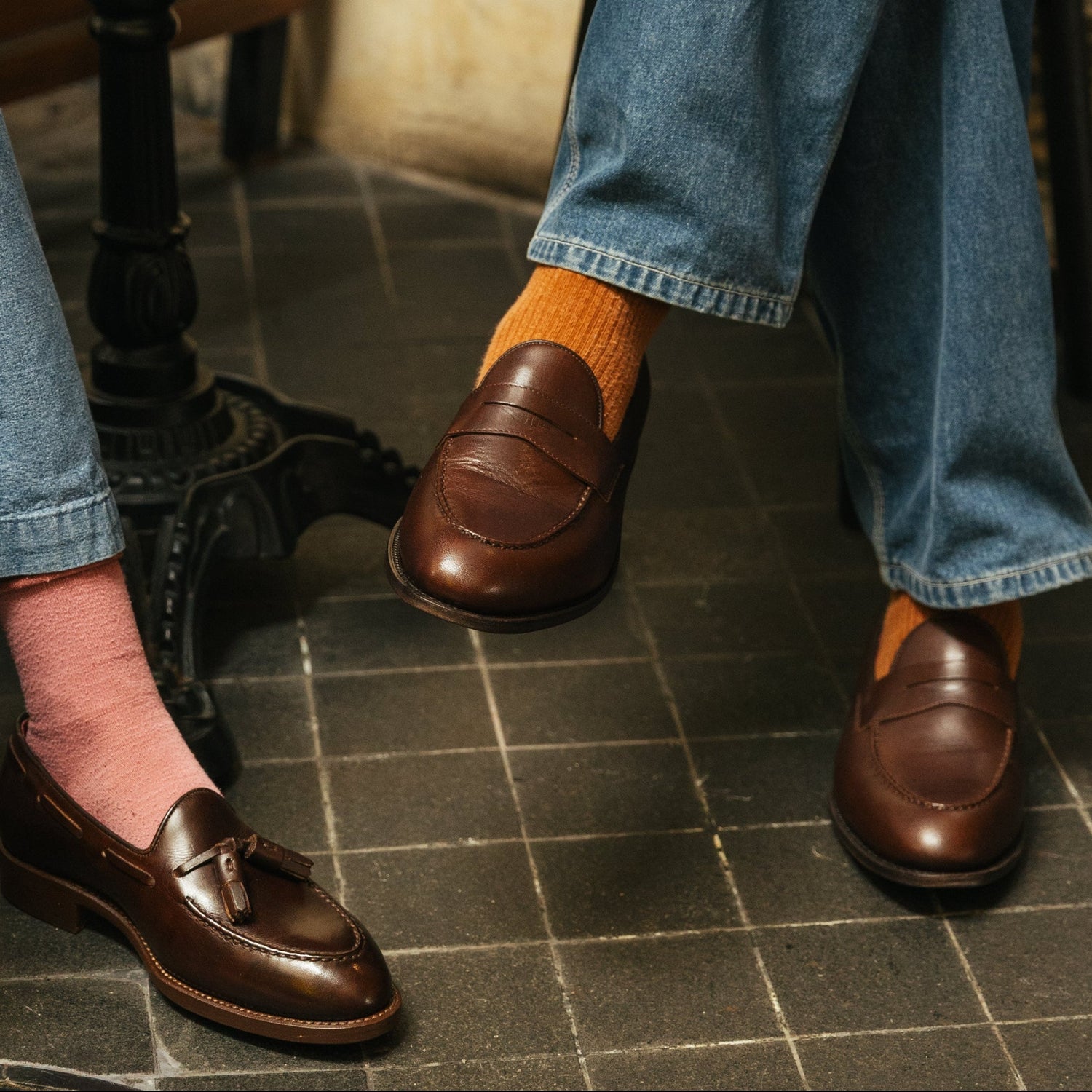 A person wearing blue jeans and brown leather penny loafers with orange socks, seated in a rustic indoor setting with a bench and tiled floor.